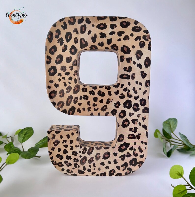 May include: A large number nine, decorated with a leopard print pattern in shades of tan, brown, and black. The number is freestanding and has a hollow center. The background is white, with green foliage accents.