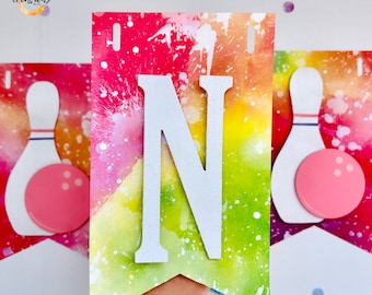 May include: A colorful banner with a large white letter "N" in the center. The banner has a watercolor effect with splashes of red, yellow, and green. Bowling pin and ball graphics are on either side.