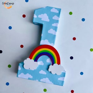 May include: A large number one birthday decoration. The number is wrapped in light blue paper with white clouds. A colorful rainbow and white clouds are attached to the bottom. Colorful confetti dots are scattered around the number.