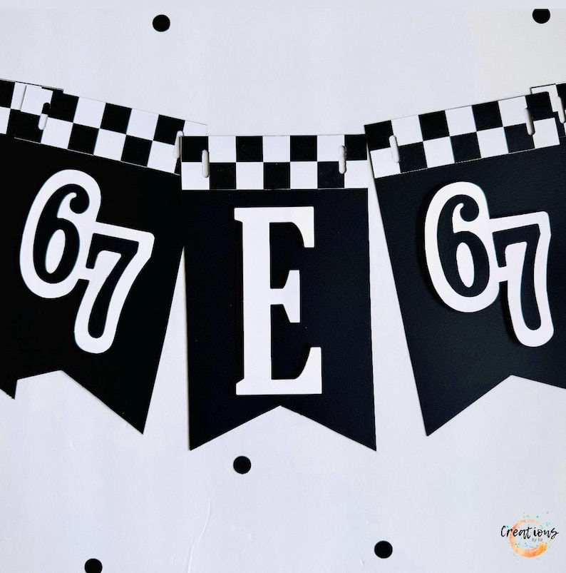May include: Black and white banner with the number 67 and the letter E. The banner has a checkered pattern and is likely for a birthday or celebration. The background is white with black polka dots.