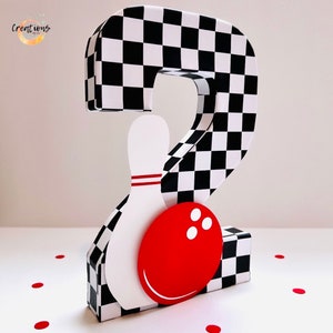 May include: A decorative number two in a bowling theme. The number is covered in a black and white checkered pattern. A white bowling pin with red stripes and a red bowling ball with white dots are attached to the number.