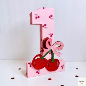 May include: A pink number one, decorated with cherry patterns and a glittery bow. Two red cherries with green stems are attached to the front. The number is made of cardboard and is a decorative item.