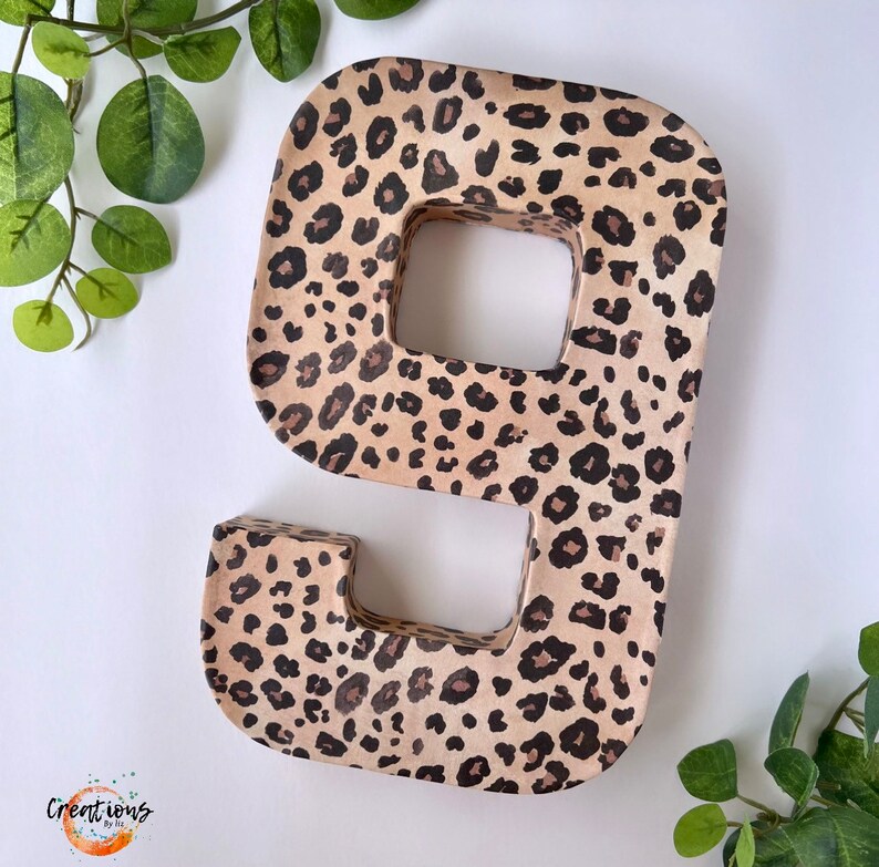 May include: A large, freestanding number nine in a leopard print pattern. The number is made of a light brown material with dark brown spots. The number is surrounded by green leaves and a white background.