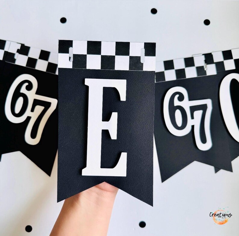 May include: Black and white banner with the letter "E" and the number "67" in a bold, white font. The banner has a checkered pattern at the top and is held up by a hand. The background is white with black polka dots.