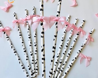 May include: White paper straws with black dalmatian spots are adorned with pink bows. The straws are arranged on a white surface, with additional bows scattered around. The image has a clean, bright aesthetic.