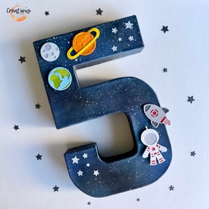 May include: A dark blue number 5, decorated with space-themed stickers. The number is speckled with white dots to resemble stars. Stickers include a moon, planet Saturn, Earth, a rocket, and an astronaut. Small star stickers are scattered around the number.