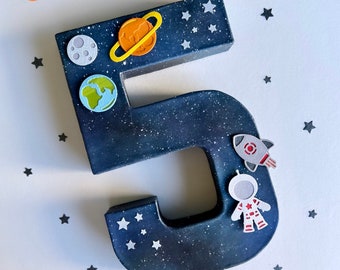 May include: A dark blue number 5, decorated with space-themed stickers. The number is speckled with white dots to resemble stars. Stickers include a moon, planet Saturn, Earth, a rocket, and an astronaut. Small star stickers are scattered around the number.