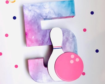 May include: A decorative number five with a bowling pin and ball. The number is painted with a watercolor effect in shades of blue, purple, and pink. The bowling pin is white with blue and red stripes, and the ball is pink.