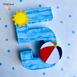 May include: A large number 5, painted blue with white wave details, decorated with a yellow sun and a beach ball. The beach ball has red, white, and navy blue sections. The number is likely for a birthday or celebration.