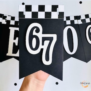 May include: Black and white banner with the number 67 in white, with a checkered pattern at the top. The banner is held by a hand. The banner is part of a set of banners with numbers and letters.