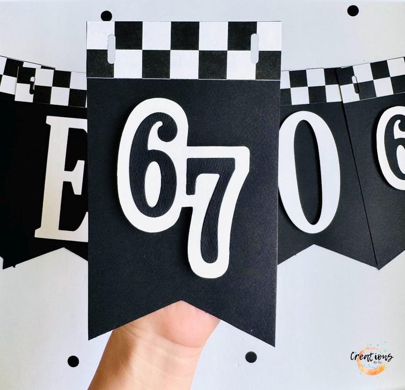 May include: Black and white banner with the number 67 in white, with a checkered pattern at the top. The banner is held by a hand. The banner is part of a set of banners with numbers and letters.
