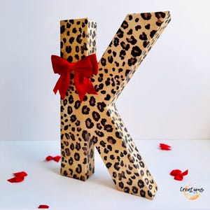 May include: A large letter "K" with a leopard print pattern. The letter is adorned with a bright red bow. Scattered around the base are red heart-shaped decorations. The letter is a decorative item.