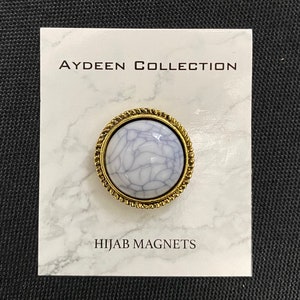 Op de afbeelding: Een witte en gouden hijabmagneet met een marmerpatroon. De magneet bevindt zich op een witte marmeren achtergrond met de tekst "AYDEEN COLLECTION" en "HIJAB MAGNETS".