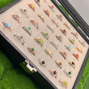 May include: A black velvet jewelry box with a silver latch, filled with wire-wrapped rings with various colored gemstones. The rings are arranged in rows and are displayed on a gray felt lining.