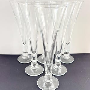 May include: Six clear, stemmed champagne flutes arranged in a semi-circle. Each glass has a slender, conical bowl and a round base. The word "MARQUIS" is printed on the base of each glass. The glasses are on a dark surface.