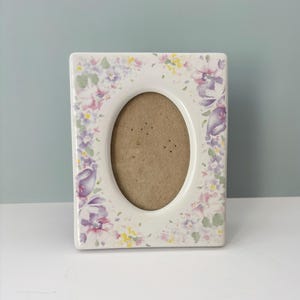 May include: A rectangular, off-white ceramic picture frame with an oval opening. The frame is decorated with a floral pattern in shades of purple, pink, and yellow. The background is a light blue.