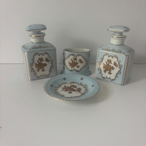May include: A set of two square light blue porcelain bottles with stoppers, a small cylindrical container, and a matching circular saucer. Each piece features a white central panel decorated with gold floral patterns and gold trim accents.