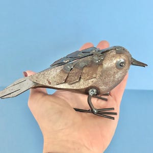 May include: A handcrafted metal bird sculpture with a textured, weathered appearance. The bird features welded metal pieces forming its body, wings, and tail. It has a small beak and is perched on thin metal legs. The sculpture is a unique piece of art.