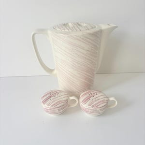 May include: A ceramic coffee pot with a matching pair of small cups, all featuring a cream-colored base with a textured, diagonal mauve-colored brushstroke pattern. The coffee pot has a tall, angular handle and a lid, while the two small cups have side handles.