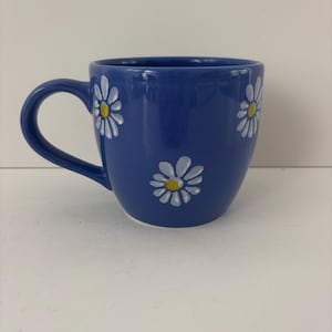 May include: A deep blue ceramic mug featuring a repeating pattern of hand-painted white daisy flowers with yellow centers. The mug has a curved handle on the left side and a smooth, glossy finish, showcasing a simple and functional design.