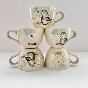 May include: A stack of six beige ceramic coffee cups. Each cup features a speckled finish and is adorned with a hand-painted design of a bird and floral elements in shades of blue, brown, and black. The cups have curved handles.