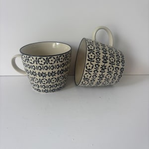 May include: Two ceramic mugs with a cream-colored base and a repeating black floral pattern. One mug stands upright while the other rests on its side. Both feature a curved handle and a dark rim, showcasing a consistent botanical design throughout.