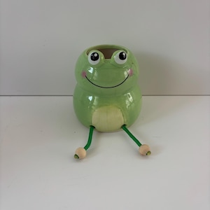 May include: A light green ceramic frog planter with a glossy finish, featuring large black and white eyes, a painted smile, pink cheeks, and a cream-colored belly. Two dangling green cord legs with small wooden beads at the ends extend outward.