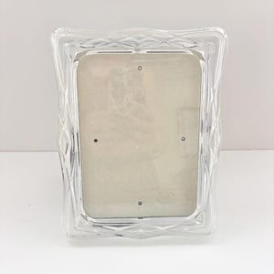 May include: Clear glass photo frame with a diamond-patterned border. The frame has a rectangular opening for a photo and is designed to stand upright. The frame is made of clear glass and has a simple, elegant design.