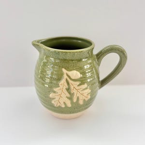 May include: A white ceramic mug with a black handle and the text "But First, Coffee" in a bold, black font. The mug is set against a blurred background, suggesting a cozy setting.