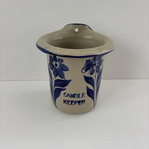 May include: A ceramic candle keeper with a beige speckled finish. It features blue floral designs and the words "CANDLE KEEPER" in blue. The keeper has a flared top with a hole for hanging.