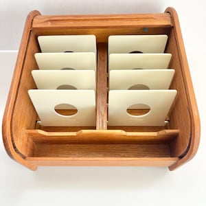 May include: A wooden record storage box with a warm, medium-brown finish. The box is divided into compartments holding off-white record sleeves. The design features a curved handle and a small shelf at the bottom, offering a vintage aesthetic.