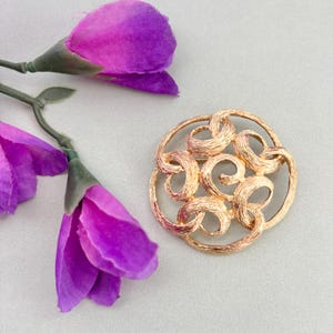 May include: A gold-toned brooch with a circular design featuring intertwined loops. The brooch is set against a light gray background, with purple fabric flowers in the upper left corner. The brooch has a textured surface.