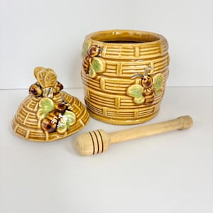 May include: A ceramic honey pot with a lid and a wooden honey dipper. The honey pot is shaped like a beehive and decorated with bees and green leaves. The lid is also beehive-shaped. The honey dipper is light brown.