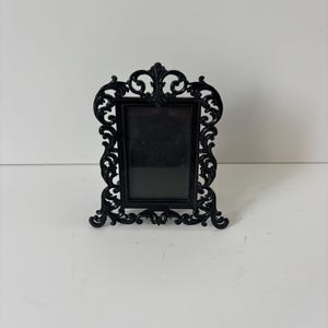 May include: A rectangular black picture frame featuring an ornate, scrollwork border with intricate swirling patterns. The frame has a vertical orientation and stands on a base with decorative feet, showcasing a glossy finish throughout the design.