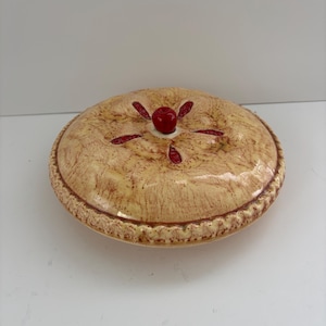 May include: A round ceramic pie dish with a lid featuring a textured, golden-brown crust finish. The lid is decorated with a central red cherry knob and four radiating red cherry-shaped cutouts, with a crimped edge detail around the perimeter.