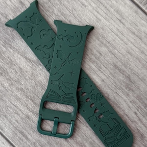 May include: A dark forest green silicone watch band featuring an engraved pattern of open books, crescent moons, stars, and swirling clouds. The band includes a matching green buckle and integrated connector ends for attachment to a digital watch.