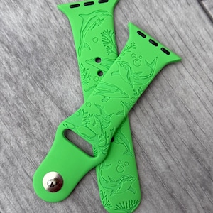 May include: Bright green watch band with a dolphin design. The band is made of a flexible material and features a detailed pattern of dolphins and bubbles. The band has a silver clasp and is designed to fit a smartwatch.