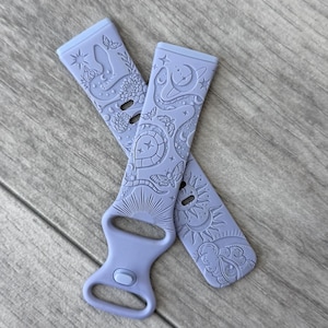 Puede incluir: Dos correas de reloj lavanda con intrincados diseños en relieve. Una correa presenta un patrón de sol, luna y estrellas, mientras que la otra muestra un tema floral y celestial. Las correas están hechas de un material flexible y están diseñadas para un reloj inteligente.