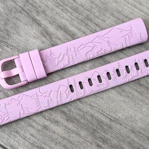 May include: A light purple watch band with a mountain and tree design. The band is made of a flexible material and has a buckle closure. The band measures approximately 9 inches long.