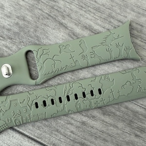 May include: Two-piece sage green watch band with a Halloween-themed design. The band features a graveyard scene with tombstones, a haunted house, and trees. The band is made of a flexible material and has a buckle closure.