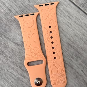 May include: Two peach-coloured watch straps with engraved designs. One strap features a moon, clouds, and an open book. The other has bats, clouds, and a stack of books. The straps are made of a flexible material.