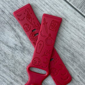 May include: Two red watch bands with a repeating pattern of spooky symbols, including bats, skulls, and potion bottles. The bands are made of a flexible material and are designed to fit a smartwatch. The bands are shown on a gray wooden surface.