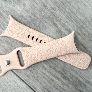 May include: Two light pink watch bands with a floral pattern. The bands are made of a soft material and are designed to fit a smartwatch. The bands are lying on a gray wooden surface.