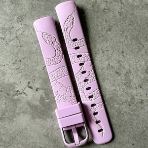 以下が含まれることがあります： ヘビのデザインが施されたピンクのシリコン製腕時計バンド2本。バンドはバックル留めです。