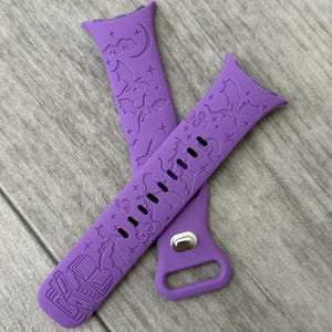Könnte beinhalten: Zwei lila Uhrenarmbänder mit einem dekorativen Design aus Fledermäusen, Sternen und einem offenen Buch. Die Armbänder bestehen aus einem flexiblen Material und haben mehrere Löcher zur Anpassung. Die Armbänder liegen auf einer grauen Holzoberfläche.