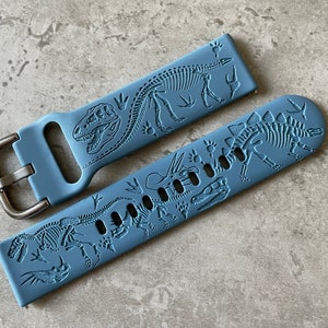 Op de afbeelding: Twee blauwe siliconen horlogebandjes met dinosaurus skeletontwerpen. De bandjes zijn gemaakt van zacht, flexibel siliconen en bevatten verschillende dinosaurus skeletten, waaronder een T-Rex, Triceratops en Stegosaurus.