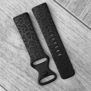 Op de afbeelding: Twee zwarte horlogebandjes met een luipaardprint. Eén bandje heeft een gesp en de andere heeft meerdere gaten voor aanpassing. De bandjes zijn gemaakt van flexibel materiaal en zijn ontworpen voor een smartwatch.