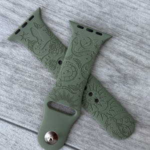 K&ouml;nnte beinhalten: Ein olivgr&uuml;nes Silikon-Uhrenarmband mit einem eingravierten Muster aus Himmels- und Naturmotiven. Das Design zeigt eine Schlange, eine Schildkr&ouml;te, eine Motte, eine Sonne, einen Mond und ein Auge. Das Band hat einen Metallstiftverschluss.