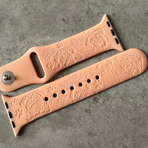 May include: A peach-coloured silicone Apple Watch band with an embossed design of pugs and flowers. The band is shown in two pieces, with the top piece showing the watch connector and the bottom piece showing the buckle.