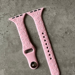 May include: Two pink silicone watch straps with a paw print design. The straps are for a smartwatch and have a silver metal connector.